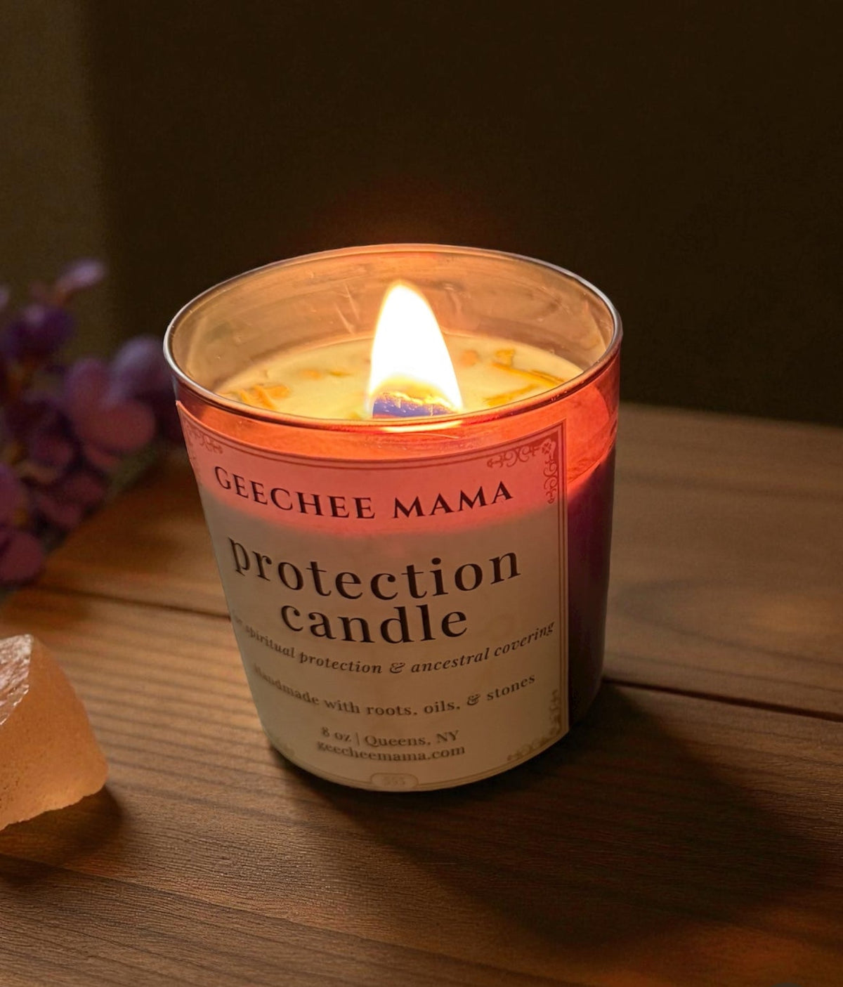 Geechee Mama ancestral protection candle burning on a wooden table beside rose quartz and purple flowers